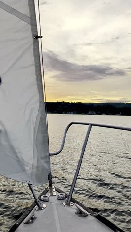Vertical view from the bow of a sailboat looking past the white jib canvas sail towards a scenic tree lined shoreline during a golden sunset on Lake Guntersville