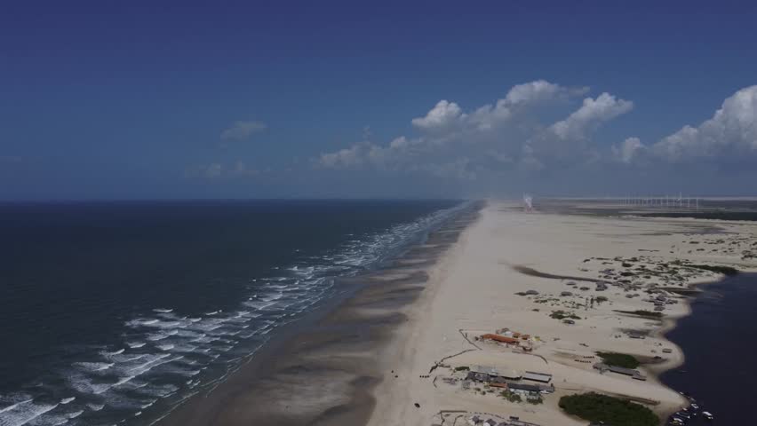 Wide aerial drone perspective showing the narrow sandy strip of Caburé surrounded by the ocean, river, and distant wind turbines in Maranhão, Brazil.