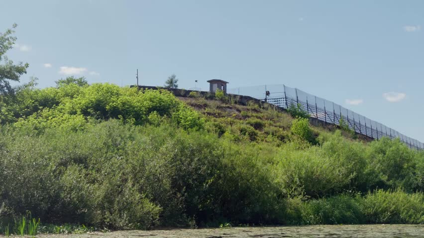 Tracking shot from river reveals high border security fence with watchtower on grassy hill under blue sky near Daugavpils, Latvia, representing European Union boundary.