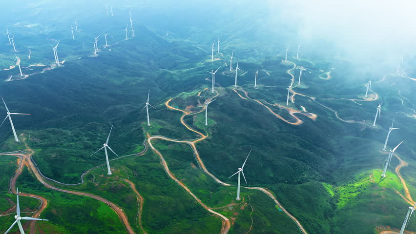Aerial view of a wind farm with massive wind turbines standing on green mountain ridges along winding roads under cloudy sky.