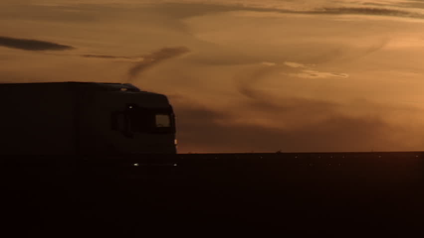 Black silhouette of truck with semi-trailers driving along road crossing sun in picturesque sunset sky with clouds in the evening twilight close-up