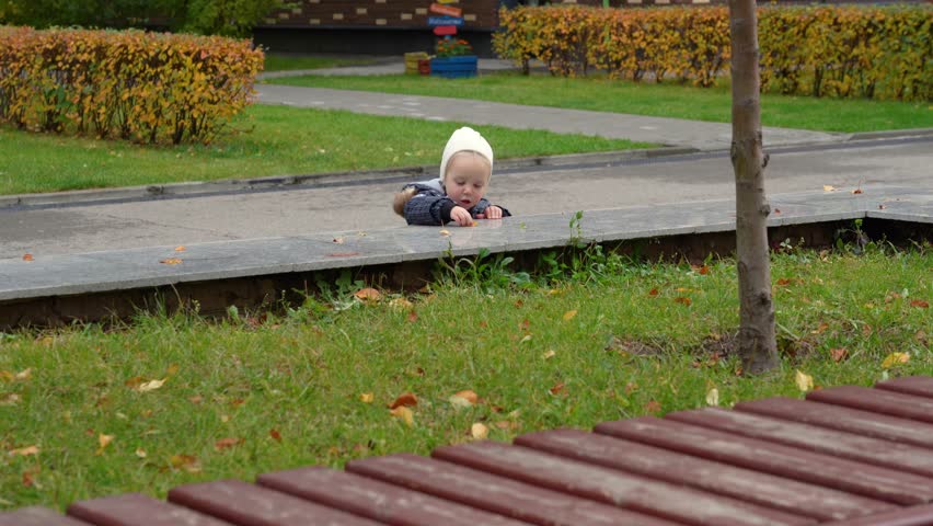 Adorable curious toddler in white beanie exploring public park courtyard, leaning on stone ledge during crisp autumn day with fallen leaf litter on green grass