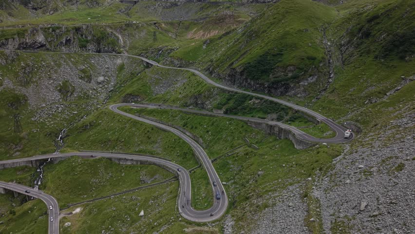 Wide static drone view showing multiple Transfăgărășan hairpin bends with continuous vehicle traffic crossing Romania