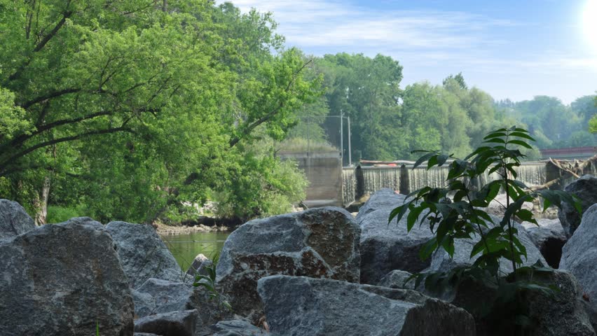 A view of a small dam on the Rock River in Watertown, Wisconsin, framed by large, textured boulders and green foliage in the foreground under a blue sky.