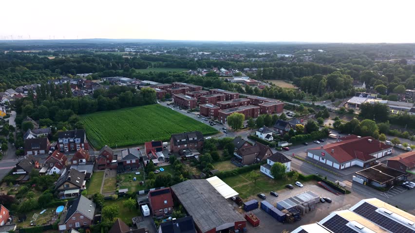 Red brick apartment blocks border green farm field beside quiet German suburb at golden sunset, tree-lined streets and red-roof houses stretch across residential district. Aerial drone forward motion