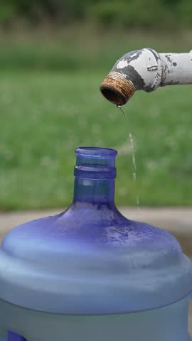 Fresh, clean water flowing from a rustic metal outdoor tap into a large blue plastic water bottle outdoors in a green park setting during a sunny summer day.