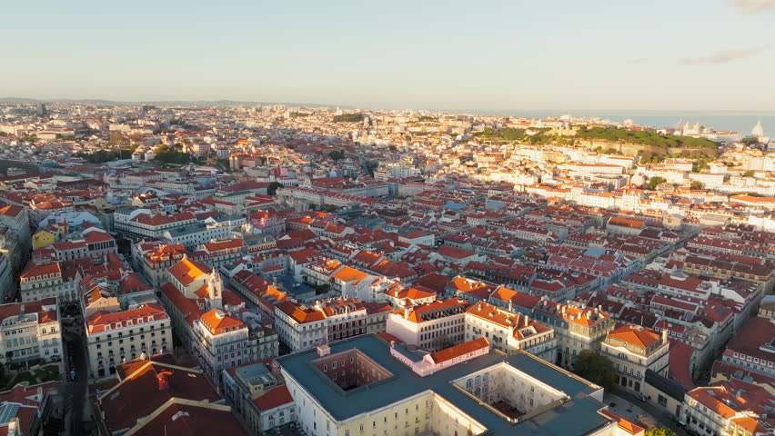 Aerial view shows Lisbon, Portugal, glowing in warm sunset light, with dense historic neighborhoods, red rooftops, elegant city blocks and the wider urban landscape stretching toward the horizon.