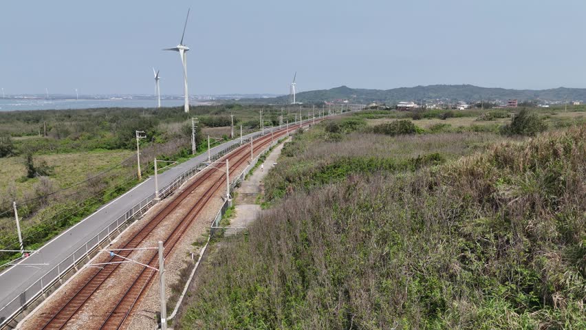 Aerial footage of an inter-city express train of EMU3000 Series dashing through green fields on a sunny summer day and giant wind turbines standing by the beach in a seaside park on the windy coast, in Houlung Township, Miaoli County, Taiwan, Asia