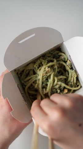 Person eating asian noodles with pesto and salmon from a takeout box using chopsticks on a white background