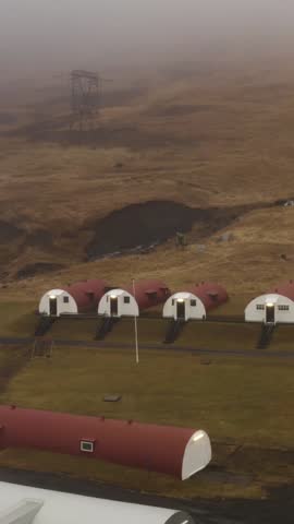 Aerial view of unique red-roofed Nissen hut accommodations in a remote, grassy valley in Hvalfjordur, Iceland. A large electricity pylon stands on a foggy hill in the background.
