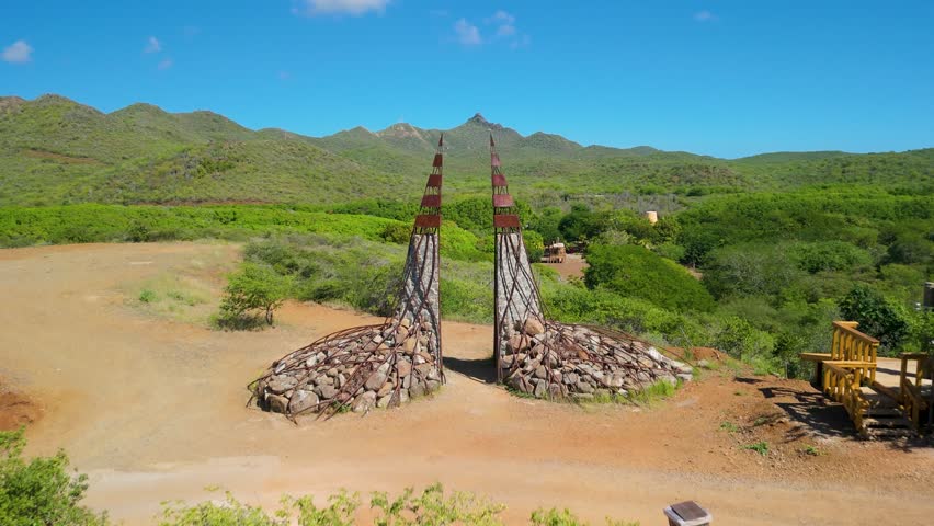 Stone and metal spires of Stairway to Heaven stand along dirt path surrounded by green shrubland and distant mountains in Curacao