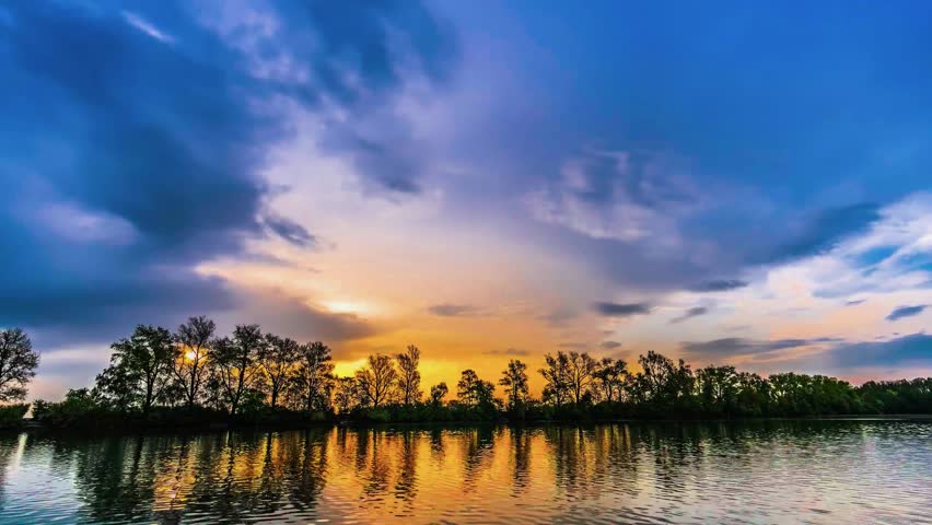A dramatic sunset time-lapse capturing turbulent stormy clouds reflecting in the calm, mirror-like waters of a tranquil lake with tree silhouettes.