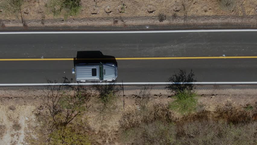 Aerial drone view of pickup truck driving along rural highway through dry landscape. Top down perspective showing vehicle road roadside vegetation and arid terrain