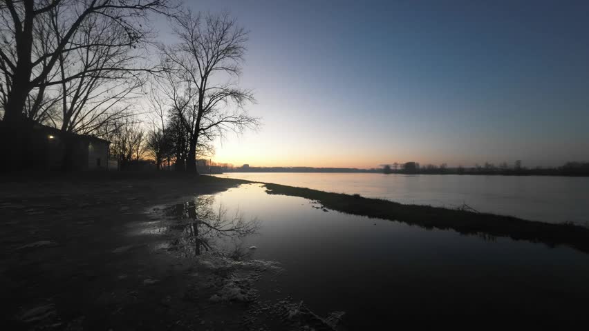 A tranquil scene unfolds as the sun begins to rise, casting a warm glow over a still river and its surrounding landscape with silhouetted trees