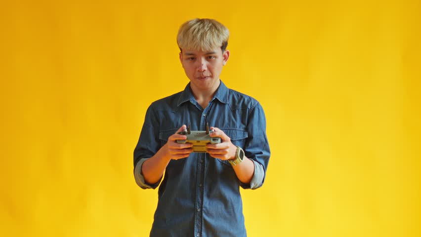 Young Asian man flying drone using remote controller against bright yellow background