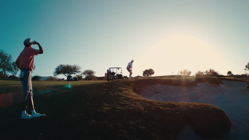 Golfer watches partner hit a pitch shot over a bunker as sunset light silhouettes the desert course in slow motion, great for golf lifestyle stories, resort ads, and travel promos.