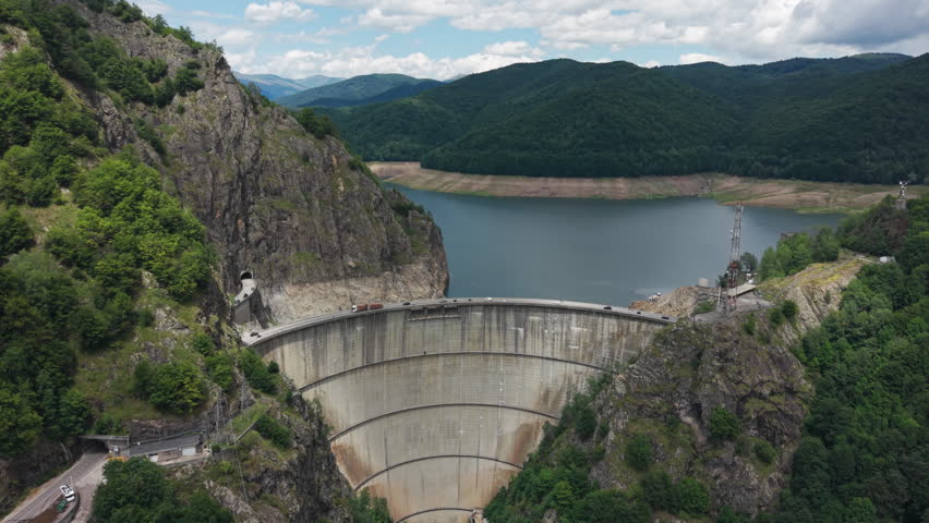 Aerial view of Vidraru Dam, reservoir, mountain tunnels, rocky cliffs, and forested hills in the Romanian Carpathians.