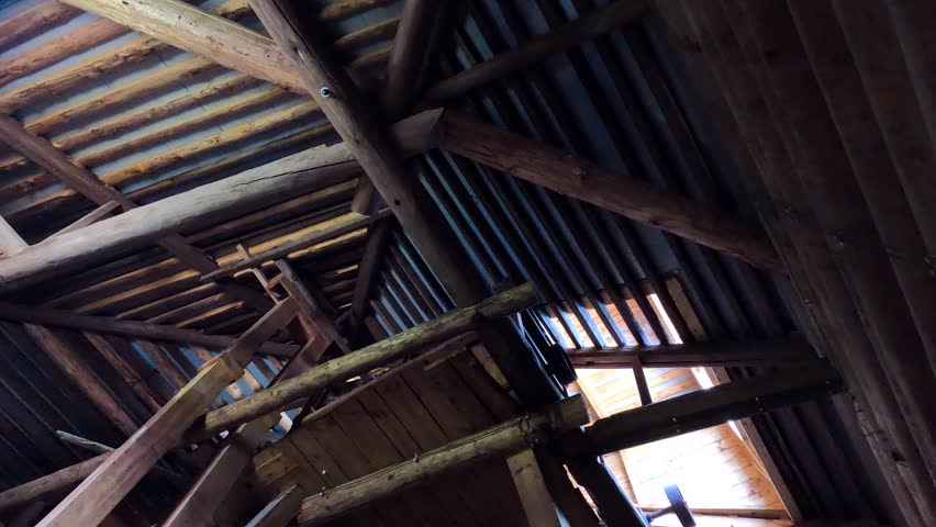 A low angle inside an old wooden mill, showing exposed beams, a staircase, and a sloping roof through which sunlight shines. Rustic wooden interior of an old mill.