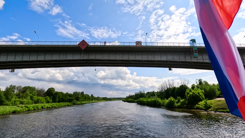 The Russian flag is flying in the foreground over the river, with a large bridge across the water behind it. The flag is installed on a ship sailing under a bridge under a blue sky.