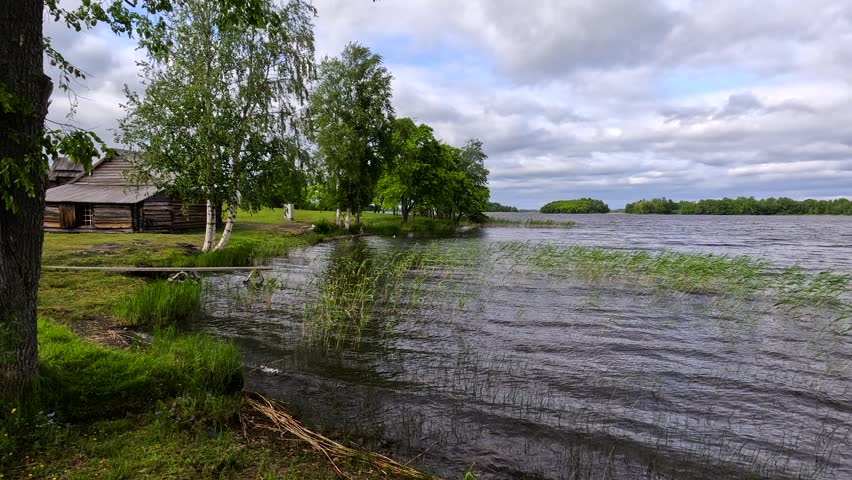 A lone wooden cabin stands by a vast lake under a dramatic cloudy sky. Reeds and grasses line the calm rippling shore.