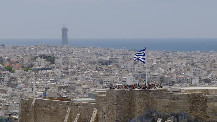 View from the ancient walls of the Acropolis over Athens, Greece, with a Greek flag flying above visitors and the city stretching to the sea.