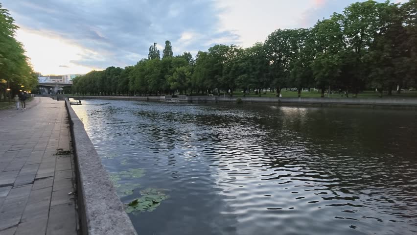 Calm riverside park features stone walkway beside quiet river, mature trees lining bank, soft reflections on water, cloudy sky above, green canopy, peaceful dusk atmosphere and gentle flow