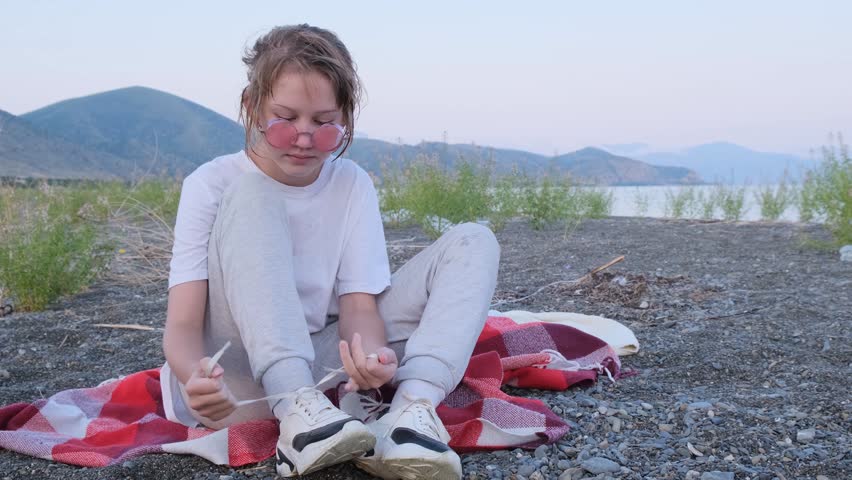 Portrait of a stylish teenage girl wearing round pink sunglasses tying shoelaces on a plaid blanket by the sea while smiling during conversation. Beautiful young girls. Wet hair.