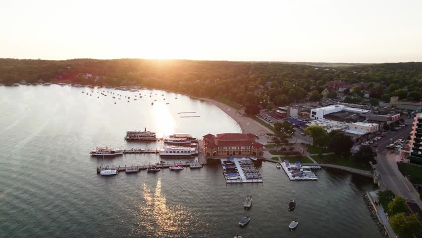 Sunset aerial view of Lake Geneva, Wisconsin, with Riviera Beach, marina docks, tour boats, anchored boats and warm golden light over the lakefront town.