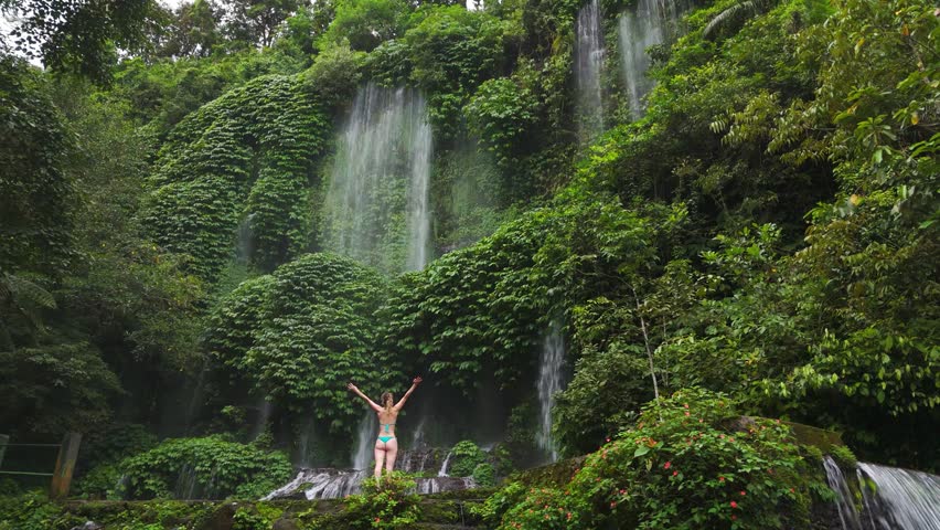 A young woman in a bikini stands in awe before the majestic Benang Kelambu waterfall in Lombok, Indonesia. Perfect for travel, wellness, and ecotourism content.