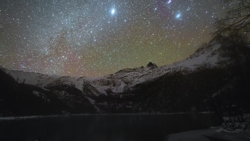 Night time-lapse footage of colorful star trails rotating across sky above snow-covered mountain landscape. Curving highway lies in mountain valley. Magnificent alpine wilderness, stunning astronomical scenery, quiet dark night in high altitude region of China.