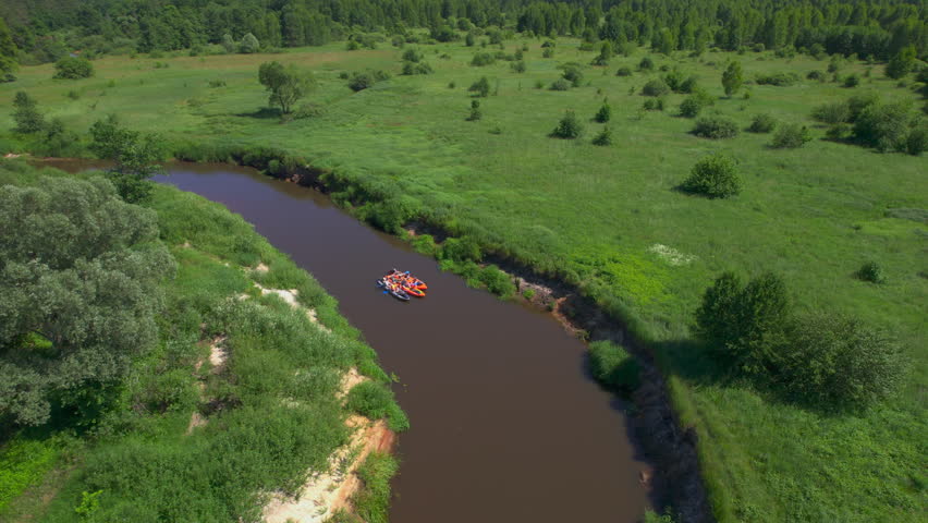 Friends kayaking through open countryside features colorful inflatable kayaks clustered on a gentle brown river bend as the drone pulls back above banks scattered trees and a summer meadow