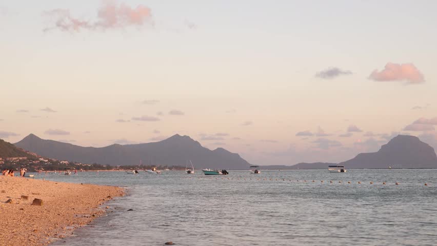 Panoramic twilight scenery overlooking calm coastal waters and distant mountain silhouettes during sunset at Flic en Flac, Mauritius, featuring scattered moored boats.