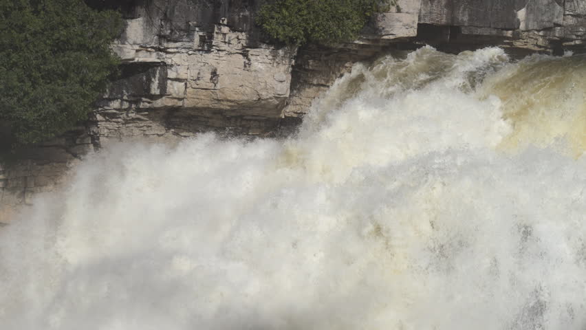 Turbulent spring water rushes over the edge of a limestone cliff at Inglis Falls. Sediment-tinted meltwater churns to white foam against the stratified rock face as cedars cling to the escarpment.
