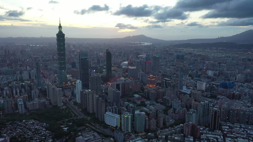 Aerial footage at dusk above Downtown Taipei, the vibrant capital city of Taiwan, with 101 Tower standing out amidst the skyscrapers in XinYi Commercial District and city lights dazzling under moody twilight sky.