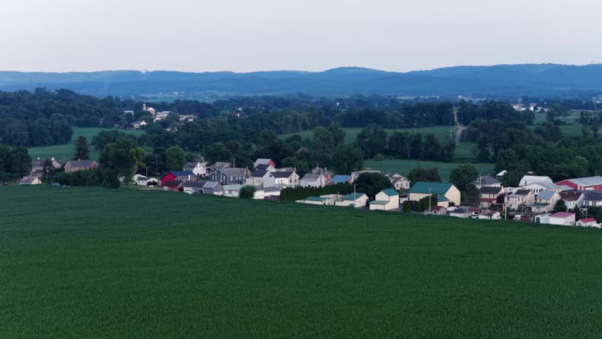 Green crop field borders small farm town lined with colorful rooftops and distant church steeple, wooded hills roll beyond quiet fields in Maxatawny, Pennsylvania, USA. Aerial drone wide shot