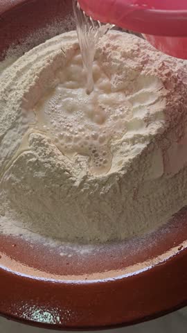 Close-up video of water being poured into flour in a traditional Moroccan clay bowl, marking the first step of homemade bread dough preparation using an authentic baking method.