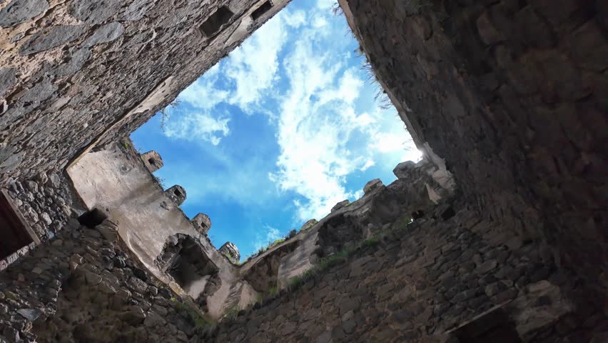 Low-angle perspective looking up at a bright blue sky with white clouds from inside the ruins of an ancient stone castle, with the sun creating a beautiful lens flare effect
