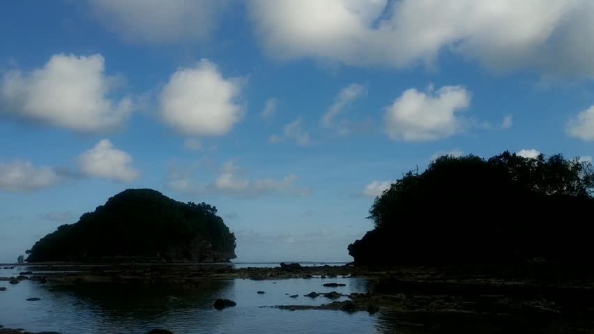 Tropical island landscape at low tide with calm blue ocean water, lush green hills, and fluffy clouds reflecting on sea surface