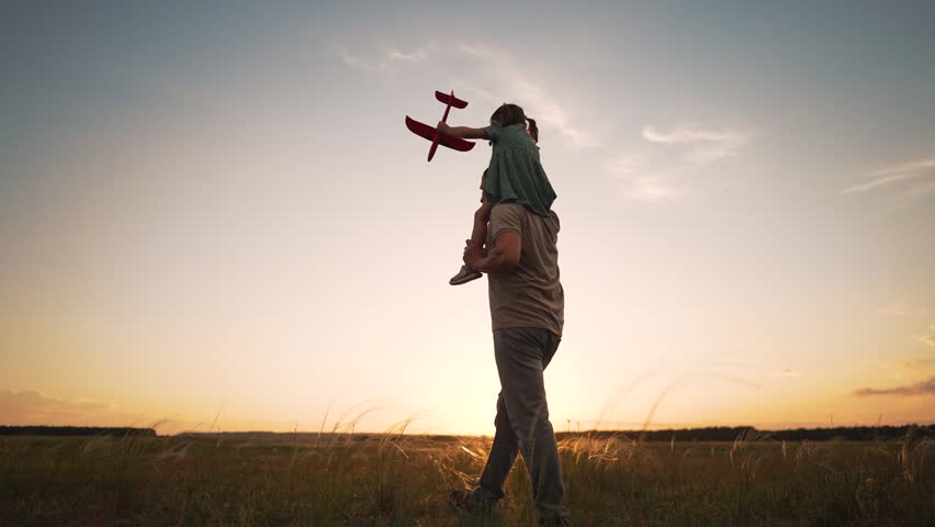Father carries child on shoulders walking through field at sunset. Happy girl holds red toy plane in air. Father and daughter play together outdoors, enjoying nature as sun sets over meadow.
