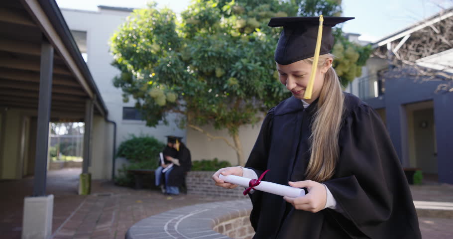 Female grads gowned in quad grad holding rolled diploma checking laughing as camera nears cheering. Campus, courtyard, outdoor, celebration, classmates, bench, tree