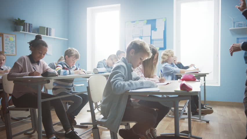 Row of Brilliant Young Kids in School Writing in Exercise Notebooks, Taking a Test. Elementary Classroom of Diverse Children Writing in Notebooks. Arc Slow Moving Camera