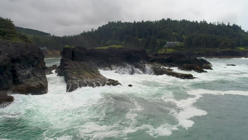 Low-flying wide-angle aerial drone shot strafing from left to right over pacific waves at Whale Cove, where surf slams a long rock barrier on the Oregon Coast south of Depoe Bay.