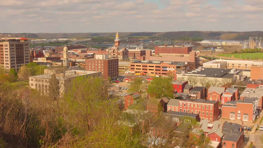 Wide panoramic aerial view of the cityscape and downtown skyline of Dubuque, Iowa, featuring historic brick buildings, urban architecture, and rolling hills in the background on a bright day.