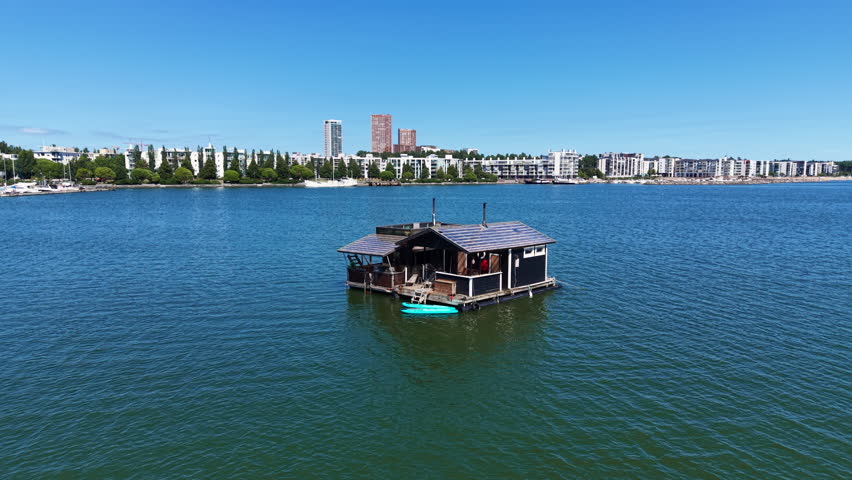 A traditional Finnish floating sauna sits on calm blue waters of Helsinki harbor with the city skyline visible in the background on a sunny summer day