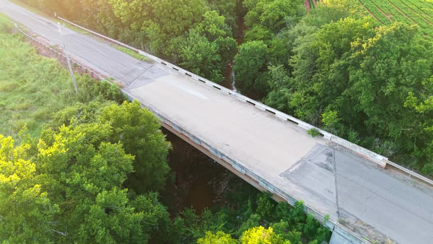 Aerial view orbiting a two-lane rural road bridge crossing over a rocky creek, surrounded by dense green forest trees and farmland in the Oklahoma countryside.