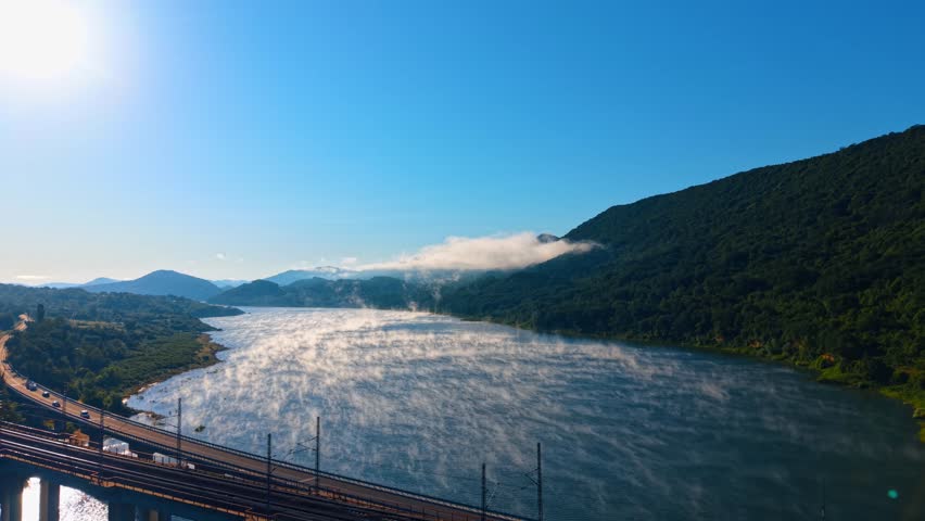 Railway bridge crossing foggy river at sunrise. Bright sunlight creates haze over calm water flowing past steep forested slopes and green fields.