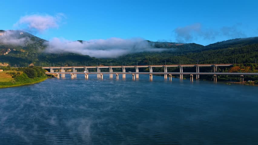 Parallel bridges spanning wide river in mountain valley. Low white clouds hang over green hills as morning mist drifts across dark blue water.
