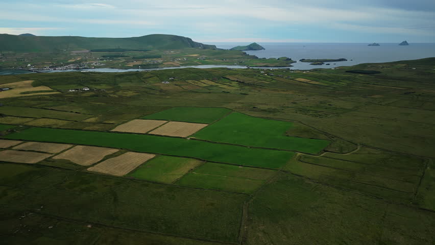 Aerial wide shot of coastal farmland showing patchwork green and brown fields with rolling hills and islands on the ocean horizon.
