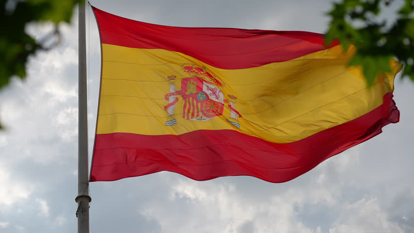 Spanish flag flying on a flagpole, with details of its coat of arms. The flag is rippling in slow motion against a cloudy sky