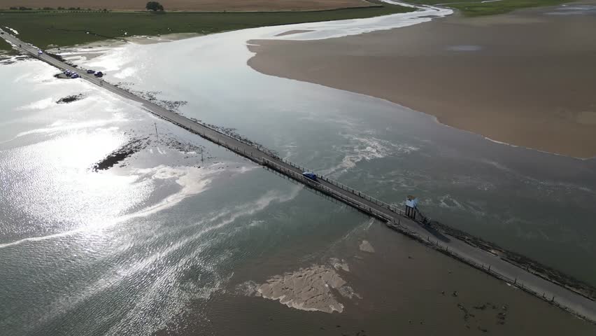 Drone footage of the Holy Island causeway featuring a blue car travelling across the tidal road. The Causeway Refuge Box stands beside the crossing as sunlight reflects off the shallow water and mudflats.